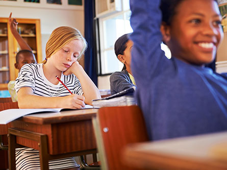 Students sit at desks in a classroom, one student raises their hand eagerly, another sits with their head resting on their head, looking bored.