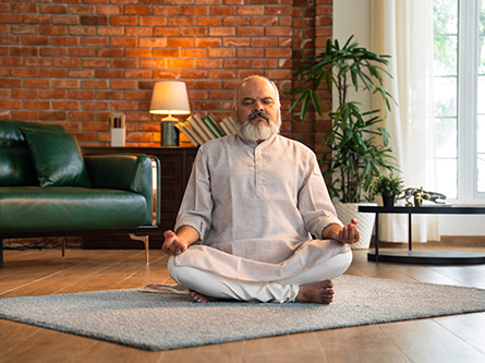 A senior man with his eyes closed sits on a carpet practicing deep breathing in a modern home.