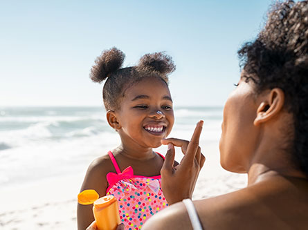 Young girl smiling as her mother puts sunscreen on her nose while at the beach to protect from skin cancer