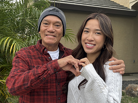 Hai and daughter Viana stand close together outdoors in front of a house and leafy plants. They are forming a heart shape with their hands.