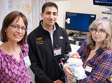 Female and male doctors in casual clothes stand next to a mother holding her infant baby in a hospital room.