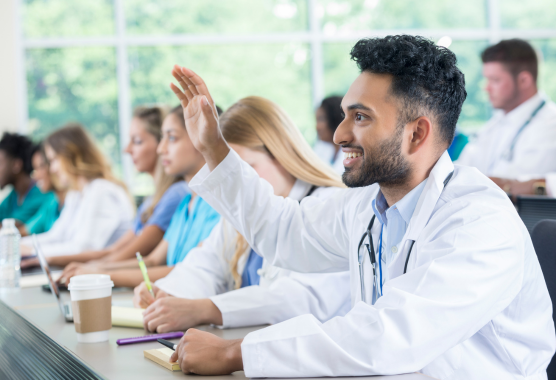 A smiling person in a white lab coat raises their hand in a classroom setting with other students in the background.