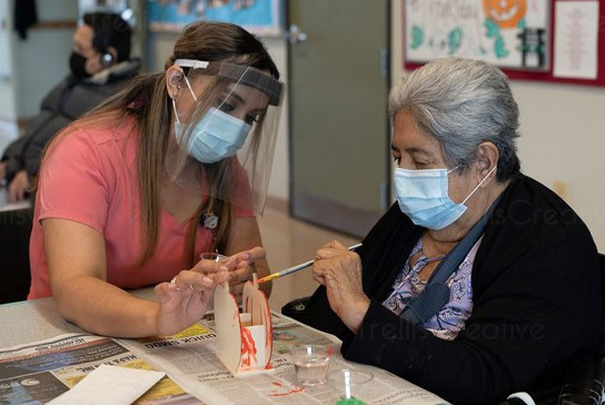 woman wearing mask and face shield helps older woman wearing mask paint a ceramic pumpkin