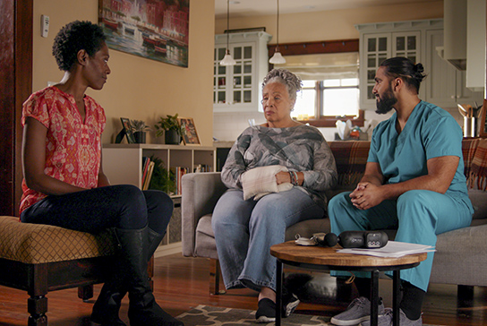younger woman in red shirt on left sits on ottoman in living room talking to older woman in middle, who holds pillow in her hands, and man in scrubs on the left looking at older woman