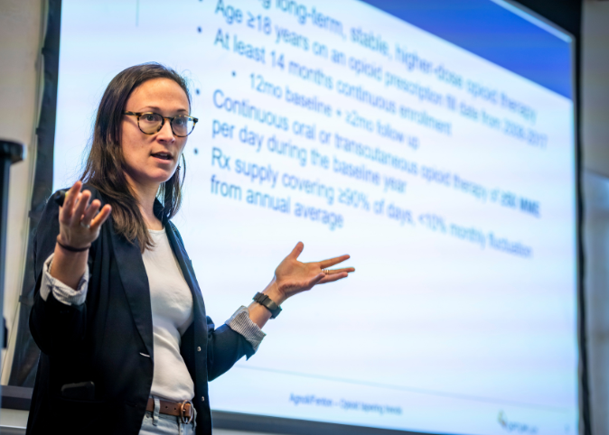 Alicia Agnoli wears a black coat and white shirt as she presents with slides on a screen in the background.