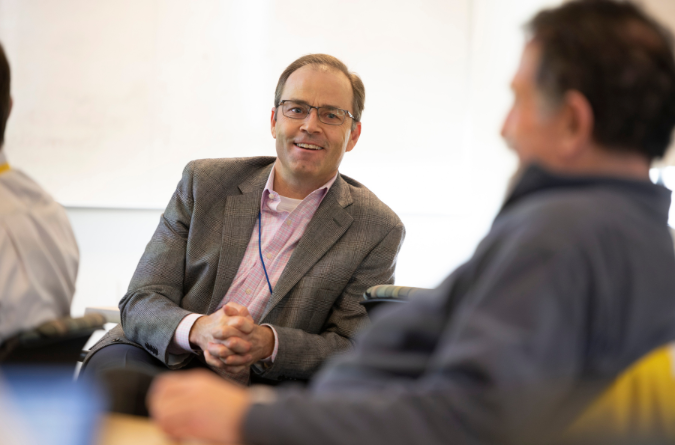 A man with short dark hair and a grey sport coat smiles while listening to man out of frame.