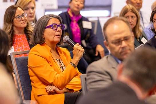 A woman in an orange jacket speaks into a microphone from the audience at the Rev PC Summit.