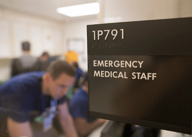 A man looks at a computer behind a glass door and sign reading 'emergency medical staff.'
