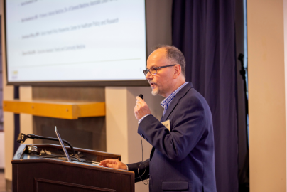 Anthony Jerant is shown from the side as he wears a dark suit and speaks into a microphone from behind a podium.