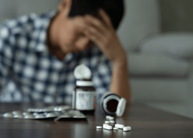 A man sits with his head in his hands behind a spilled pill bottle. 