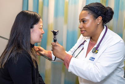 A woman physician in a white medical coat examines a woman patient in a long sleeve shirt.