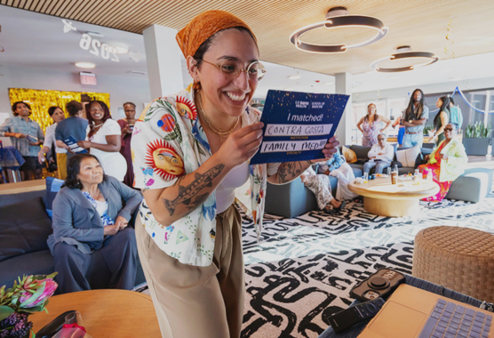 A medical student holds an "I Matched: Contra Costa Family Medicine" placard for her classmates to see in a video meeting.