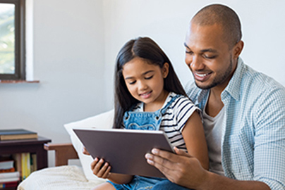 A father and daughter sitting on a bed reading from a computer tablet.