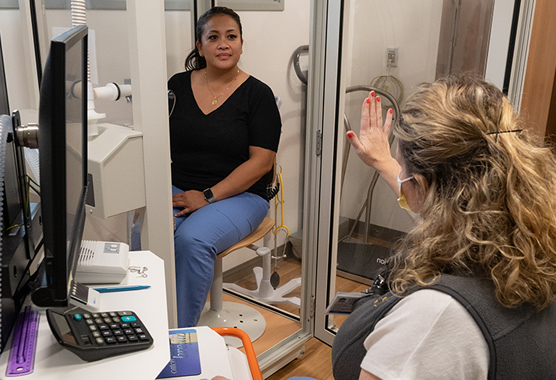 A health care worker performing a lung plethysmography test. Another woman is sitting in a telephone booth like box that can measure breathing.