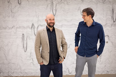 Two men stand in front of a wall conversing