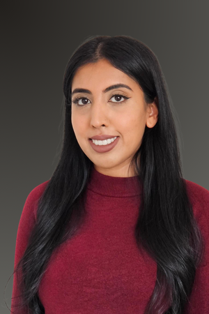 Professional photo of Ashley Balley smiling at camera. Woman is wearing a dark red top against a gray background. 