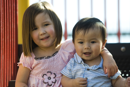 Very young older sister sits with arm around infant brother while sitting on jungle gym.