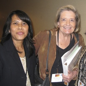 Two women standing side by side, smiling at the camera; one is holding a binder and wearing a name badge