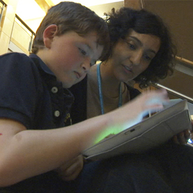 A child uses a tablet while an adult sits beside the child, looking at the screen
