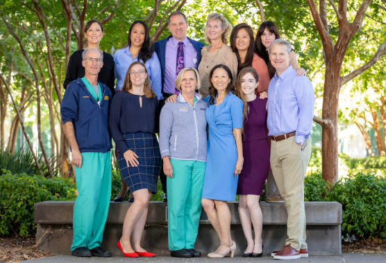 A group photo of doctors stand next to an outdoor bench and in-front of trees and bushes.