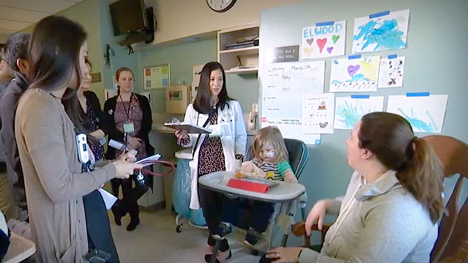 Pediatric residents listen to doctor discuss with pediatric patient and mother who are sitting down while all others are standing.