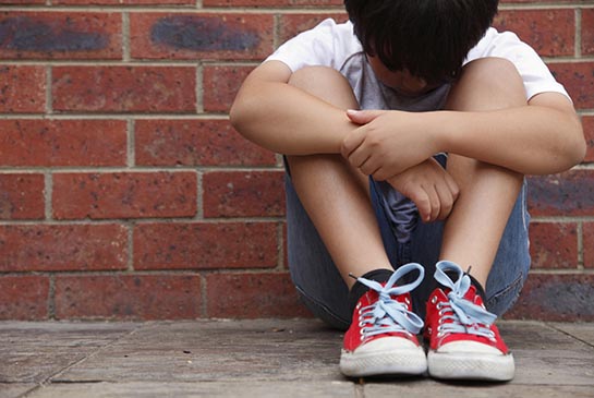 Small child sits against an outside brick wall with head low.