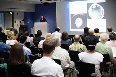 Presenter at podium displays medical photos on large screen while audience watches.