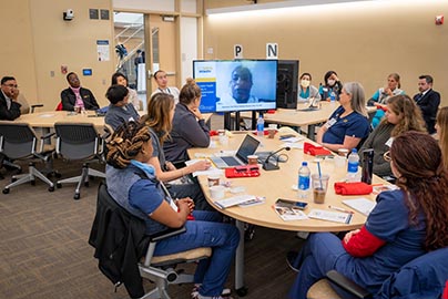 Faculty in tele-conference sitting around table, watching speaker on monitor.