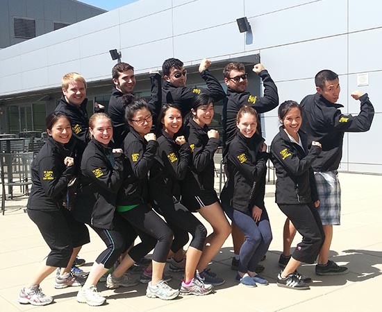 Twelve residents in black jackets and black pants posing for photo outside of hospital classroom, flexing their biceps in fun.