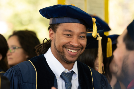 UC Davis Ph.D. graduate in graduation robe and hat, smiling.