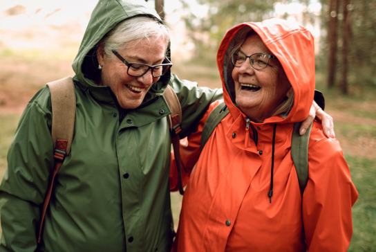 Two older women with their arms around each other laughing as they hike through the woods.