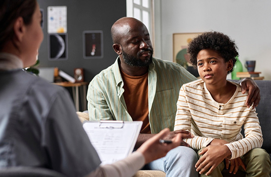 Young boy sitting with father and listening to a psychiatrist holding a clipboard