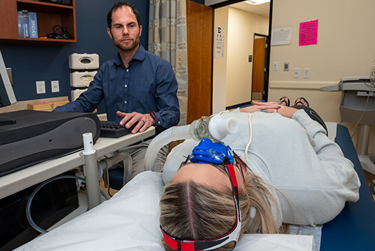 A woman lying down wearing a breathing mask, connected to an analytic machine.