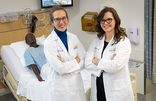 Piri Ackerman-Barger, left, and Jessica Draughon Moret, right, stand with arms crossed in front of manikin lying in hospital bed