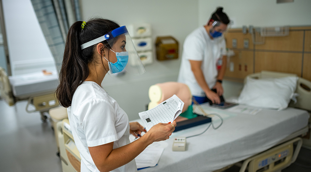 Nursing student wearing mask and faces hield looks right and points gloved finger while other nursing student in background works on manikin