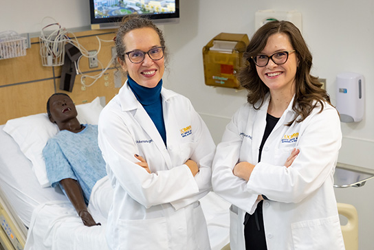 Piri Ackerman-Barger, left, and Jessica Draughon Moret, right, stand with arms crossed in front of manikin lying in hospital bed