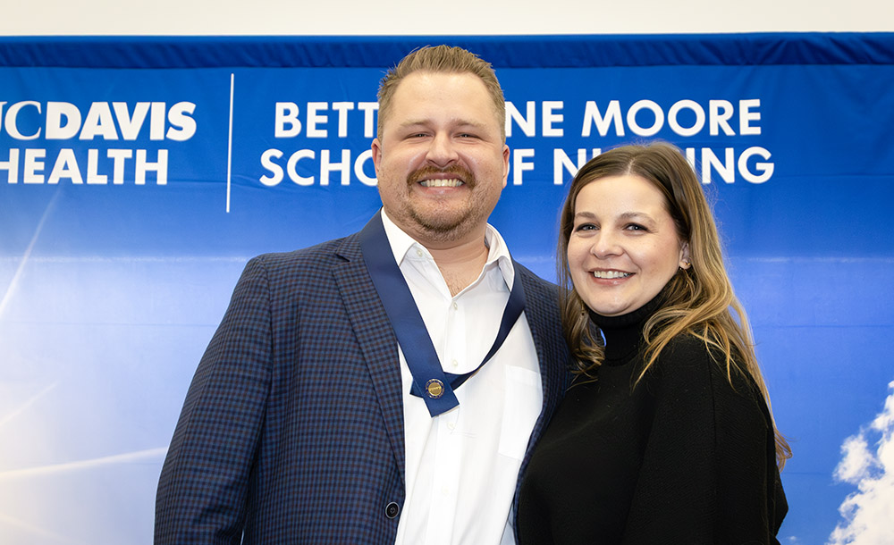 Two people smile and pose together in front of a Betty Irene Moore School of Nursing at UC Davis backdrop during a recognition or graduation event.