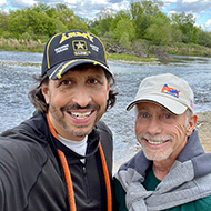 Two men looking at the camera with a hill and sky behind them